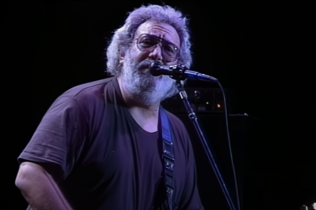 A man with a beard and sunglasses is singing into a microphone while playing an electric guitar. He is wearing a dark t-shirt, and the background is black, making the colors and details of the singer prominent.