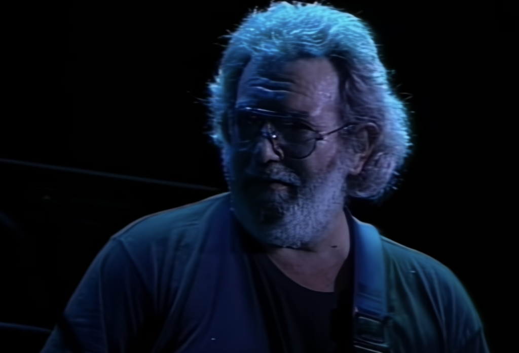 A man with curly, greying hair and a beard, wearing glasses and a dark t-shirt, stands on a dimly lit stage holding a guitar. The background is dark, highlighting his face and upper body.