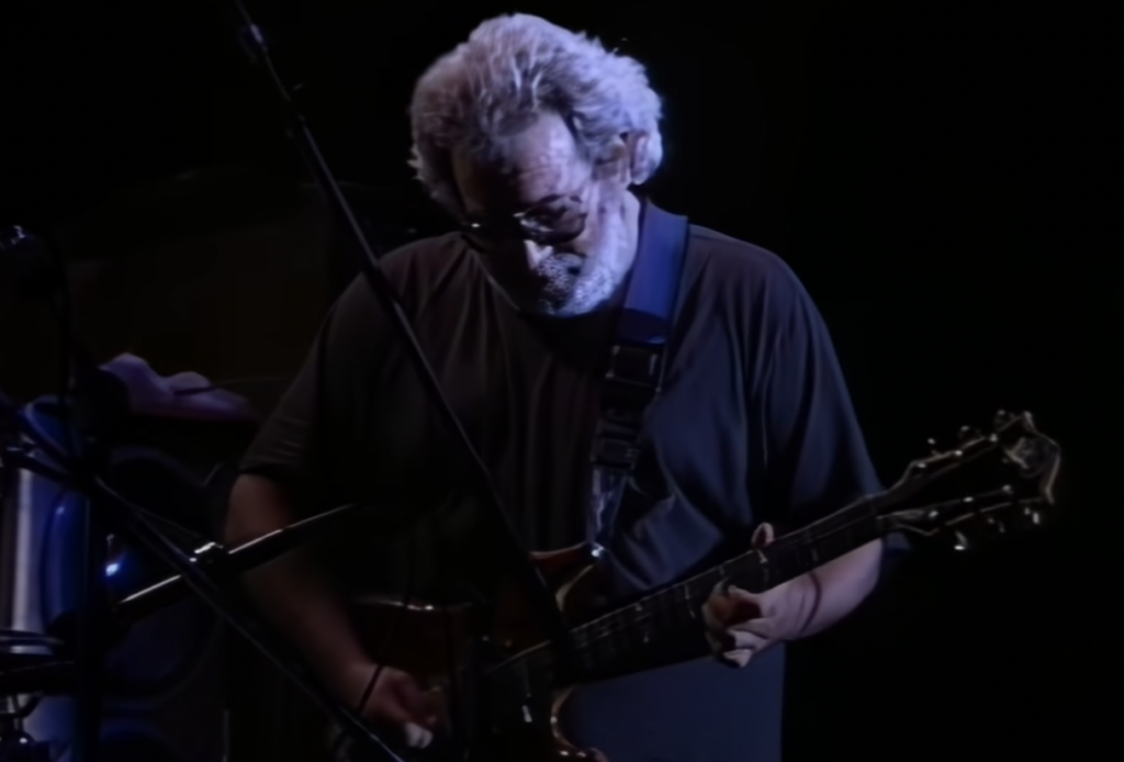 An individual with gray hair and a beard is playing an electric guitar on stage under dim lighting. Microphone stands and other stage equipment are visible around him. He is wearing glasses and a dark t-shirt.