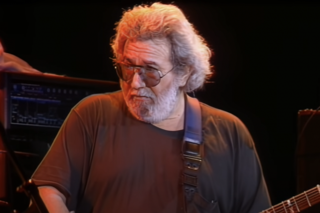 A man with gray curly hair, a white beard, and sunglasses is playing an electric guitar on stage. He is wearing a dark t-shirt, and the background shows musical equipment and dim lighting, suggesting a live performance.