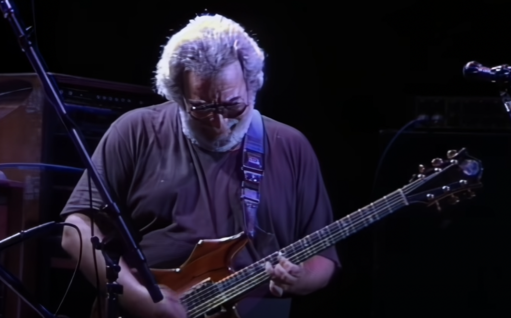 A person with gray curly hair, beard, and wearing sunglasses, is playing an electric guitar on stage. They are dressed in a dark t-shirt and appear focused on their performance. Microphones and musical equipment are visible in the background.