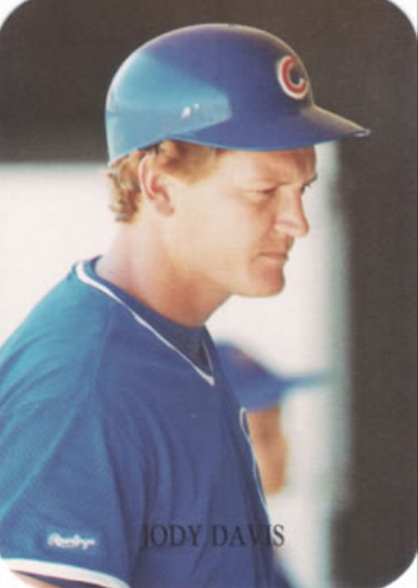 A baseball player wearing a blue Chicago Cubs uniform and a blue batting helmet looks downward. The name "Jody Davis" is written at the bottom of the image. The background is blurred, with faint figures visible.