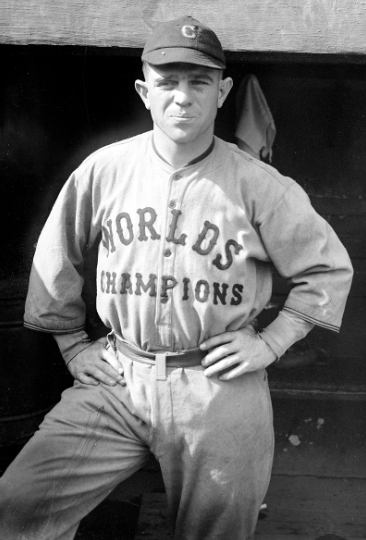 Black and white photo of a baseball player wearing a cap with the letter "C" and a jersey that reads "WORLD'S CHAMPIONS." He is standing with his hands on his hips, looking directly at the camera, and a dugout is visible in the background.