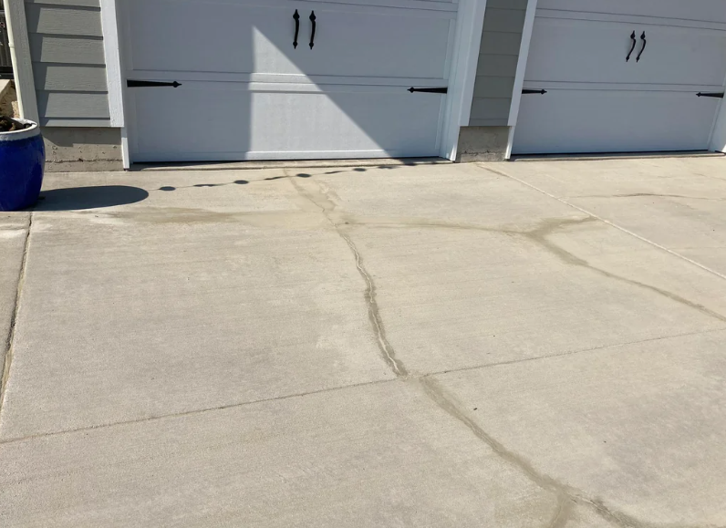 A concrete driveway with noticeable cracks runs up to a double garage with white doors. A large blue ceramic pot is on the left side of the driveway. The garage doors have black handles and hinges as decorative elements.