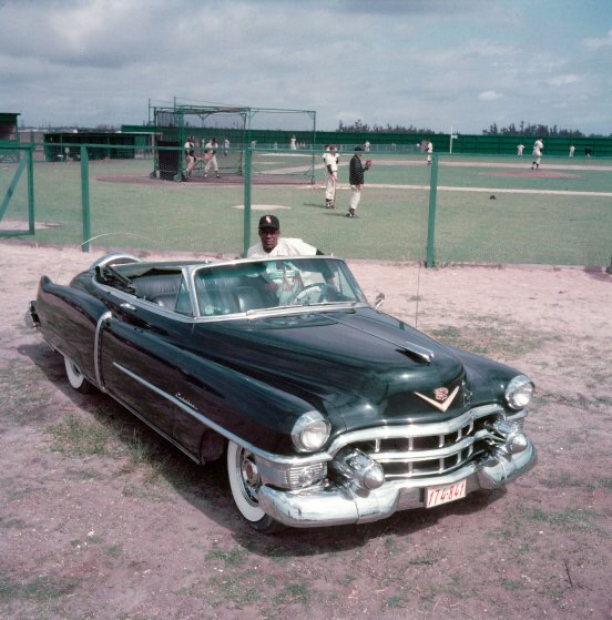 A man sits in a vintage dark green convertible parked on a grassy field near a baseball field. In the background, players practice on a baseball diamond enclosed by a green fence. The sky is partly cloudy.