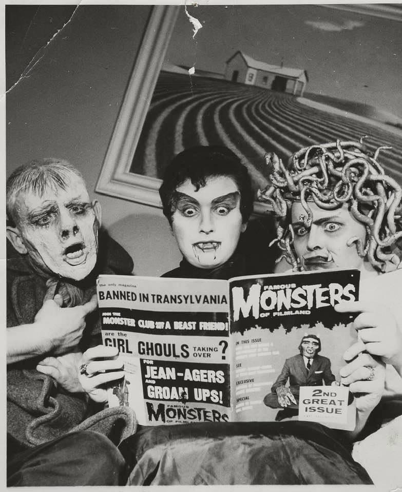 A black and white image shows three people dressed as various monsters engrossed in a "Famous Monsters" magazine. The middle person’s wide-eyed, shocked expression is exaggerated, while the others react dramatically. A rural painting hangs in the background.