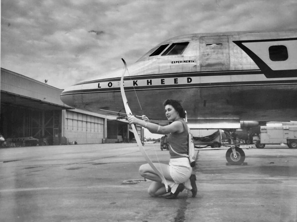 A woman kneels on the ground, aiming a bow and arrow, in front of a Lockheed airplane parked on a tarmac. The airplane has "Lockheed Experimental" written on its front. The scene appears to be an old black-and-white photograph, with a dramatic sky overhead.