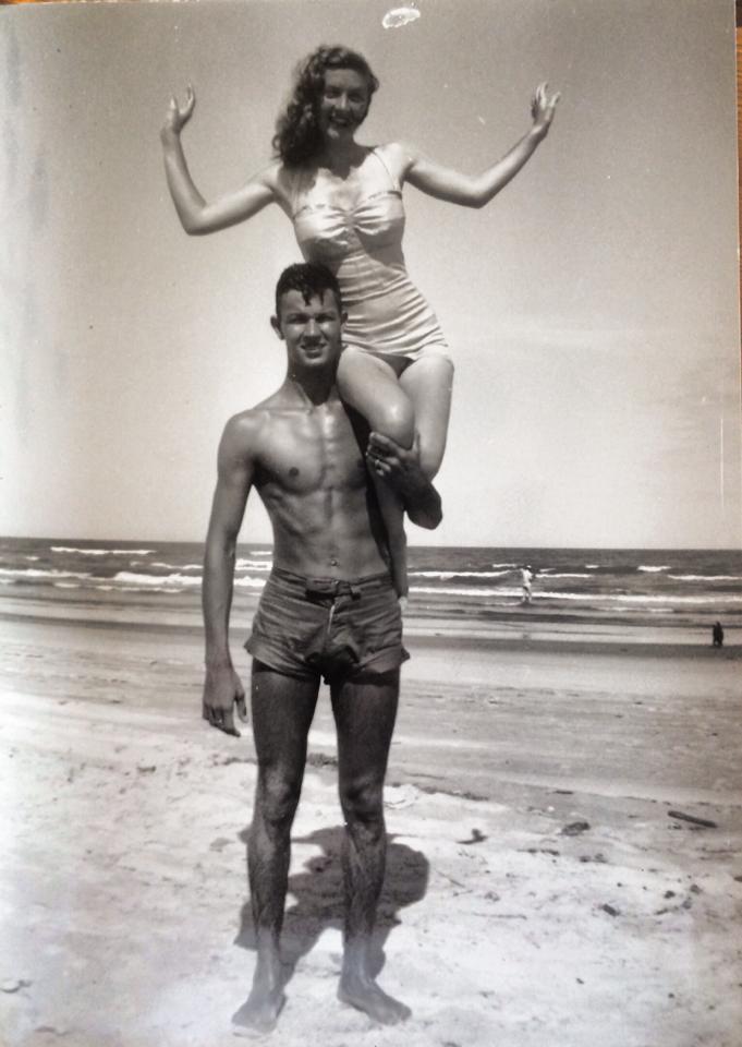 A black-and-white photo of a man and a woman at the beach. The man, wearing swim trunks, is standing on the sand while the woman, wearing a swimsuit, is sitting on his shoulders with her arms raised. The ocean and a few beachgoers are visible in the background.