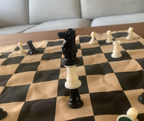 A close-up of a chessboard with pieces arranged unusually. A black knight is balancing on top of a white queen, which in turn is sitting on a black rook. Other pieces are scattered around the board. The background shows a gray sofa.