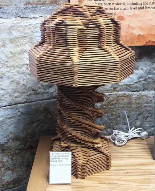 A uniquely designed wooden lamp with an intricate spiral pattern displayed on a wooden table against a stone wall. The lamp has a geometric, multi-layered structure and an electrical cord is visible next to it. A small informational plaque is placed in front of the lamp.