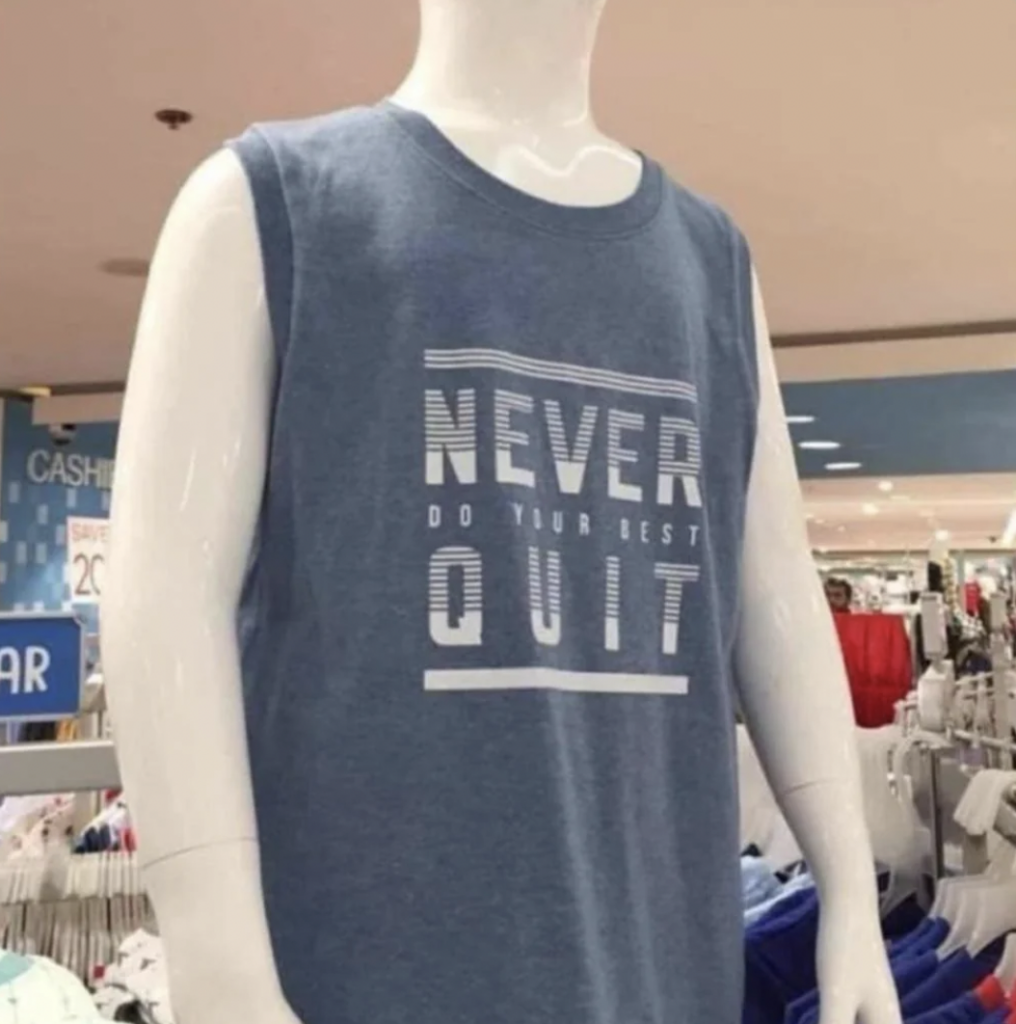 A store mannequin is wearing a sleeveless blue shirt with white text that reads "NEVER QUIT" in bold letters and "DO YOUR BEST" in a smaller font beneath it. The background shows various store aisles and clothing displays.