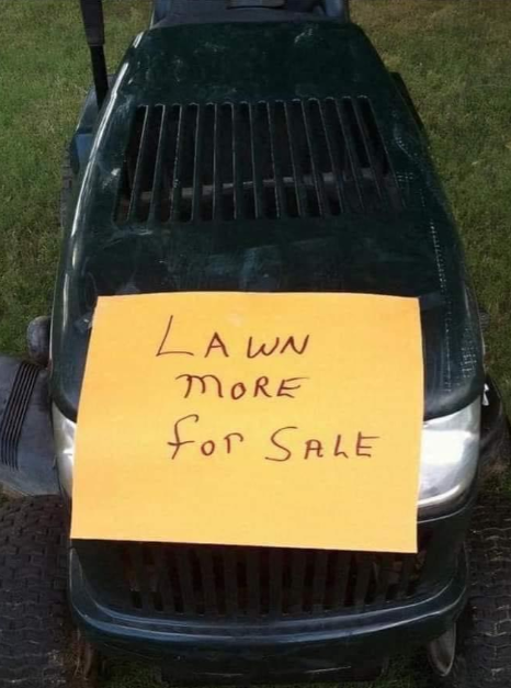 A dark green lawnmower is on grass with an orange sign taped to the front. The sign reads, "Lawn More For Sale" in handwritten letters.