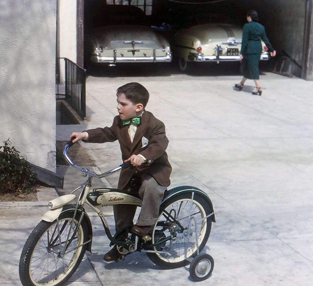 A young boy wearing a brown suit with a green bowtie rides a tricycle with training wheels near a garage with old cars parked inside. In the background, a person in a green outfit with high heels walks away from the garage.