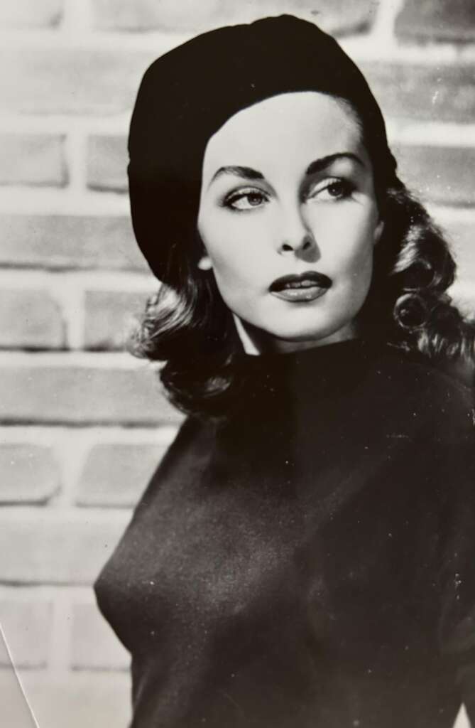 Black and white photo of a woman with wavy hair, wearing a dark beret and a matching high-neck top. She is looking off to the side with a thoughtful expression. A brick wall serves as the background.