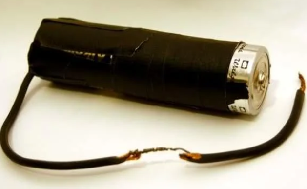 A makeshift electronic device consisting of a battery wrapped in black tape with two wires extending from it, having exposed copper ends connected together. The image suggests a basic, improvised battery pack or electrical circuit.