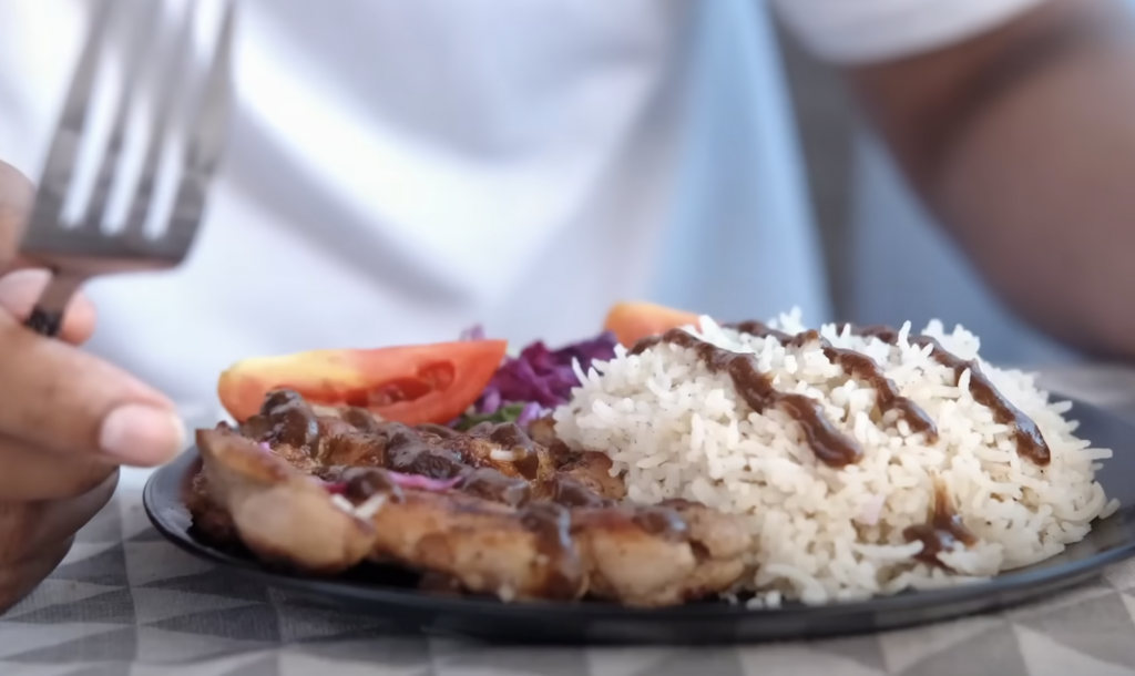 A person holding a fork is about to eat food served on a black plate. The plate contains grilled chicken with sauce, a serving of white rice, and sliced tomatoes with shredded purple cabbage. The person is wearing a white shirt.