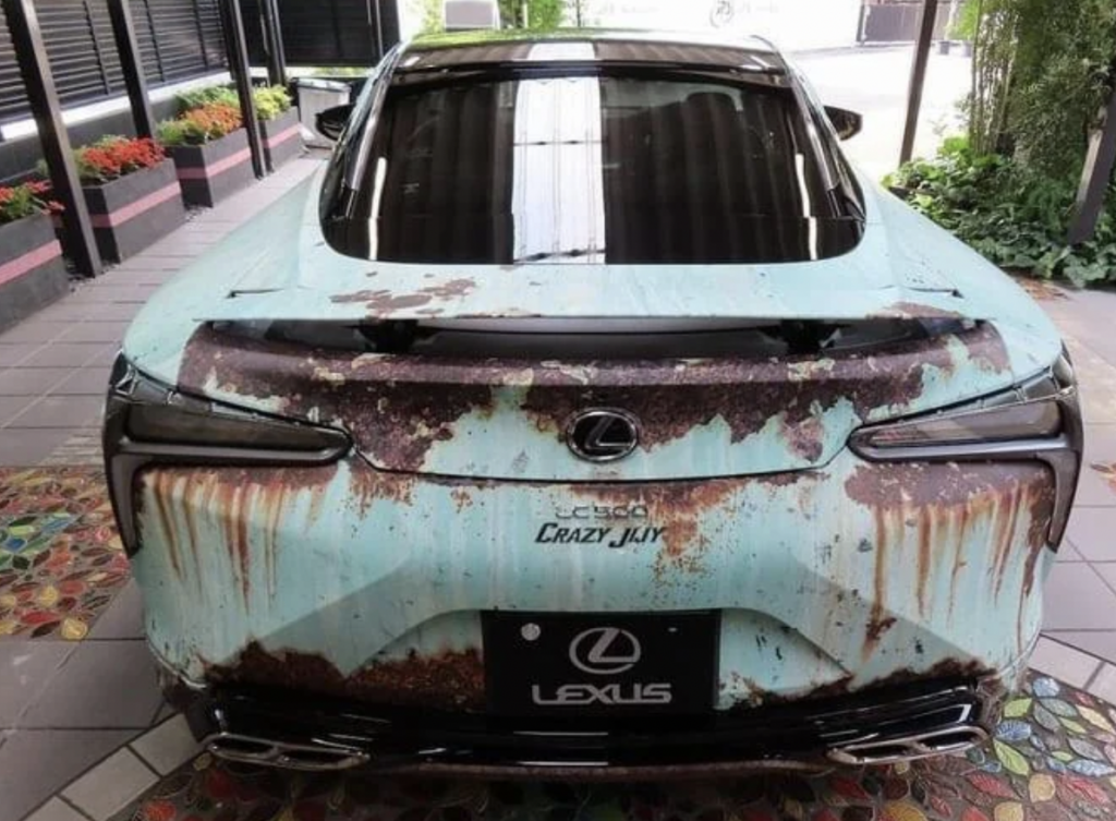 A luxury Lexus car with a heavily rusted and weathered rear exterior sits in an indoor space. The car's paint is peeling, revealing large rust patches across the back. Flowers in planters and greenery are visible in the background.