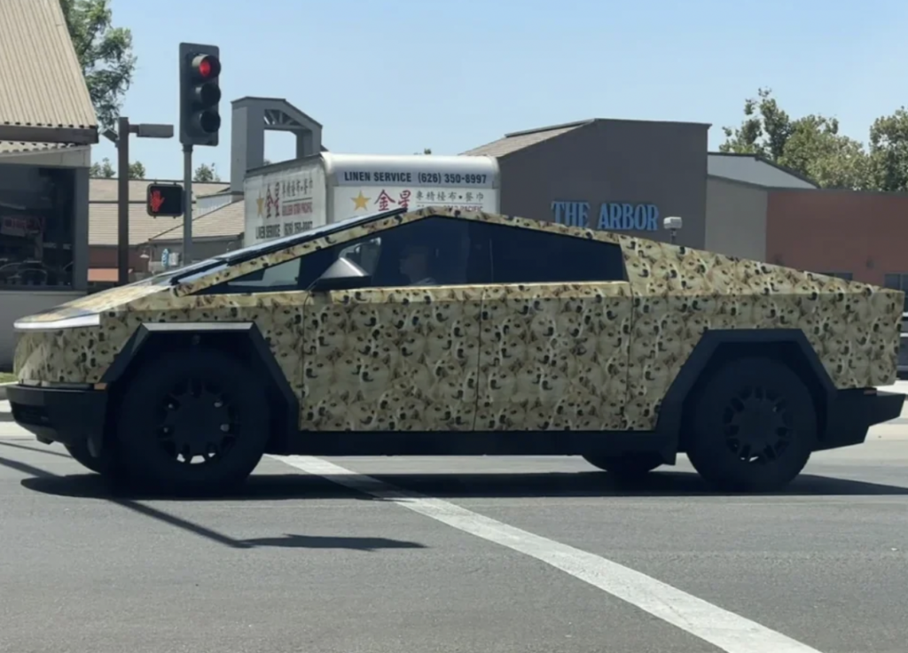 A futuristic, angular vehicle with a camouflage paint design waits at an intersection. The car has a unique, geometric shape and high ground clearance, resembling a concept or special utility vehicle. Trees and buildings are visible in the background.