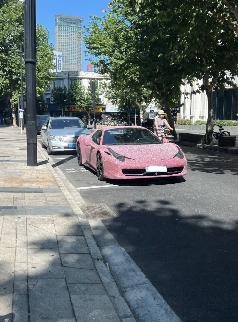 A pink sports car with a sparkling exterior is parked on the side of a city street, partially in front of a silver car. Trees line the sidewalk, and a tall building is visible in the background. A person wearing a hat walks on the sidewalk nearby.