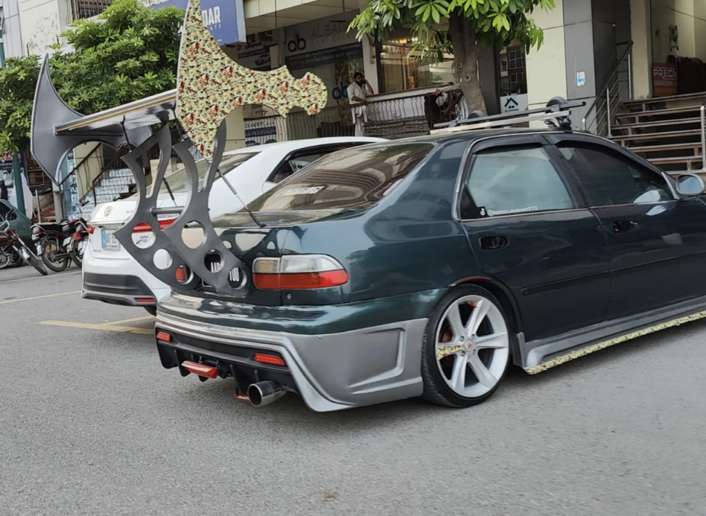 A modified dark green sedan is parked on a street, featuring a large, uniquely shaped rear spoiler with a floral pattern. The car has custom rims, an aftermarket exhaust, and other noticeable modifications. Several other vehicles and trees are visible in the background.