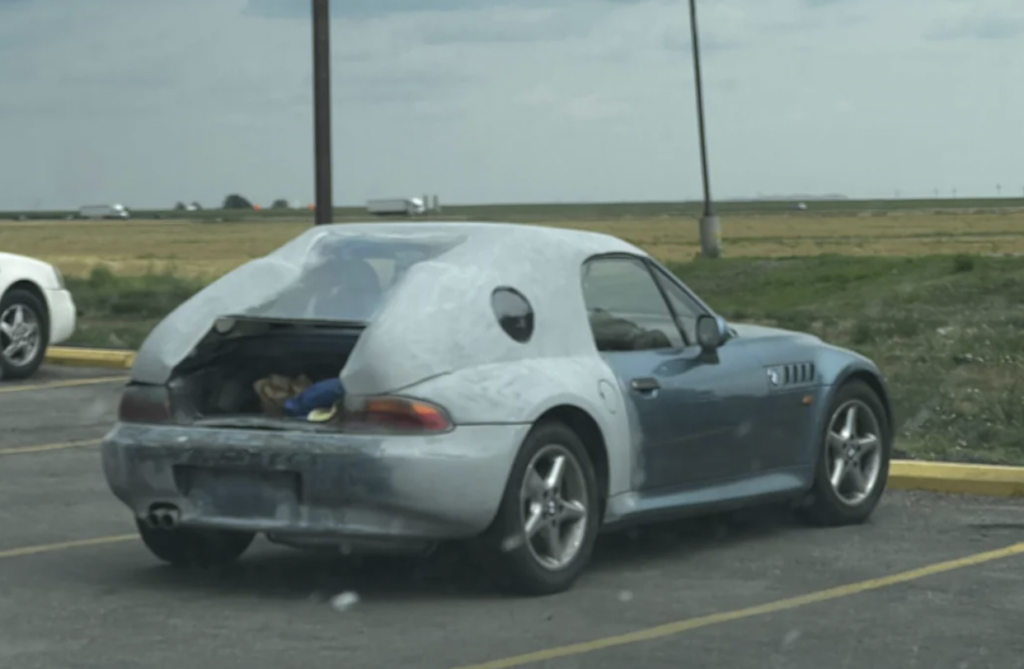 A silver-blue convertible car parked in a lot with a makeshift gray hardtop cover that extends over the rear, including the trunk area. The cover has an uneven surface and a circular cutout window on the side. The background features a grassy field and distant horizon.