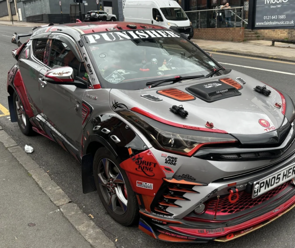 A highly modified Toyota CH-R is parked on a street. The car features an elaborate red and silver paint job with decals, a custom hood, additional lights, and various body modifications including fender flares and a front splitter. The windshield reads "Punisher.