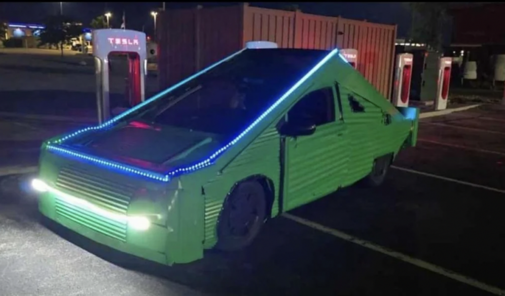 A green, unusually designed car with a triangular, corrugated metal exterior and blue LED lights on the edges is parked at a Tesla charging station at night. The car's headlights are on, illuminating the dark area around it.