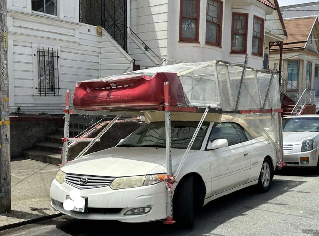 A white car is parked on a residential street with a large, unconventional red tent-like structure on its roof. The structure is secured with metal poles and appears homemade. The surrounding houses have steps leading up to their entrances.