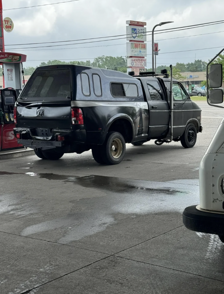 A black pickup truck with a camper shell is parked at a gas station. The truck has extended side mirrors, and the rear wheels appear modified or in repair. The gas station has a red fuel pump and signs in the background. The sky is cloudy.