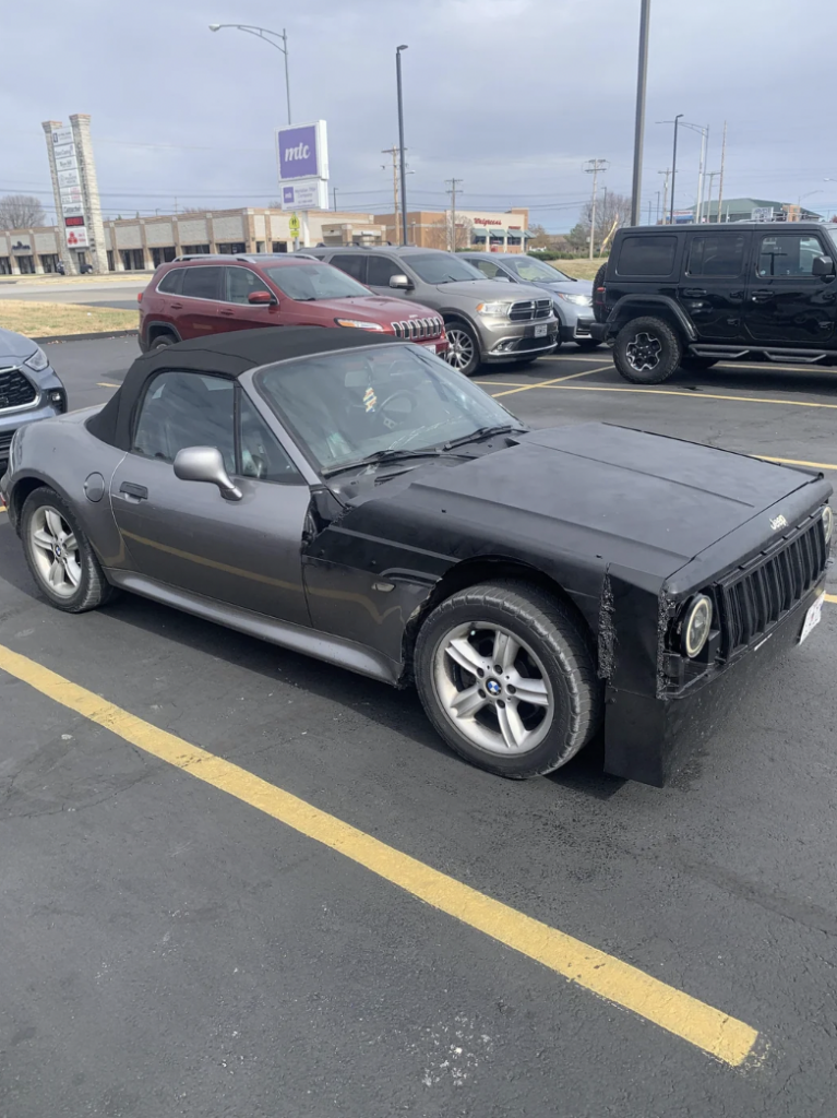 A black and silver convertible car is parked in a parking lot. The front of the car has been modified to resemble a Jeep, with a distinctive seven-slot grille. Several other vehicles and buildings are visible in the background.