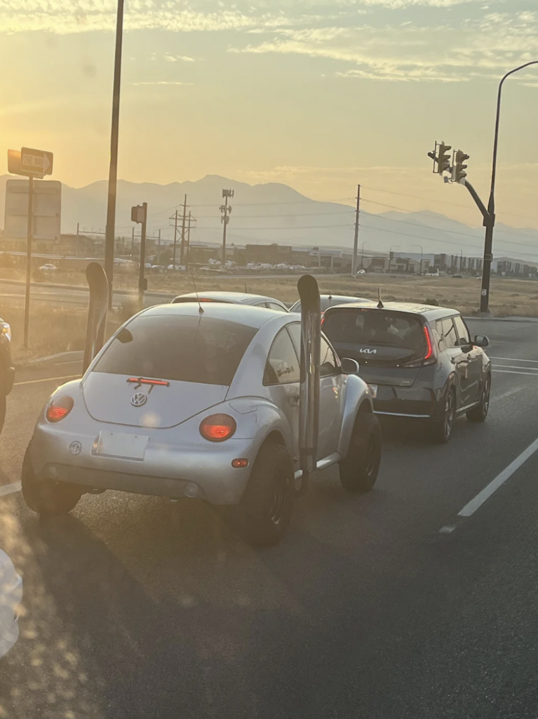 A modified Volkswagen Beetle with large exhaust stacks and oversized tires is stopped at a traffic light. A silver SUV is positioned ahead, while the sun sets over distant mountains, casting a warm glow across the scene.