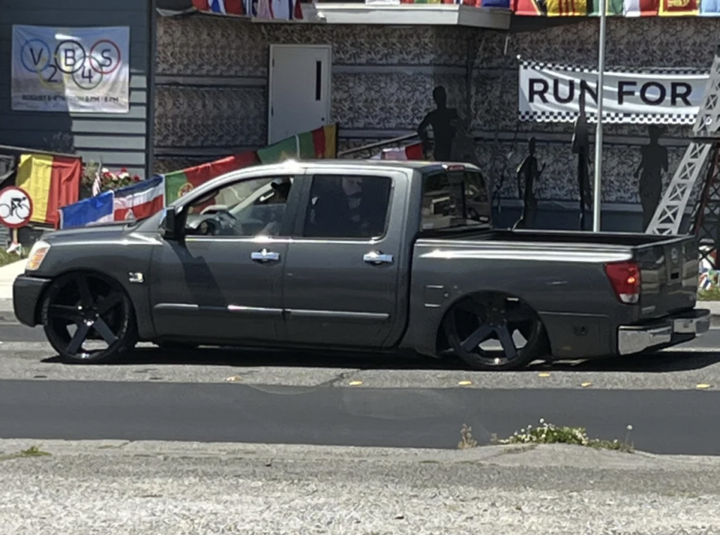 A low-riding, modified grey pickup truck with large black rims is parked on a street. Several colorful flags and banners are visible in the background, and there's a "Run For" sign partially visible on a building. The street appears sunny and clear.