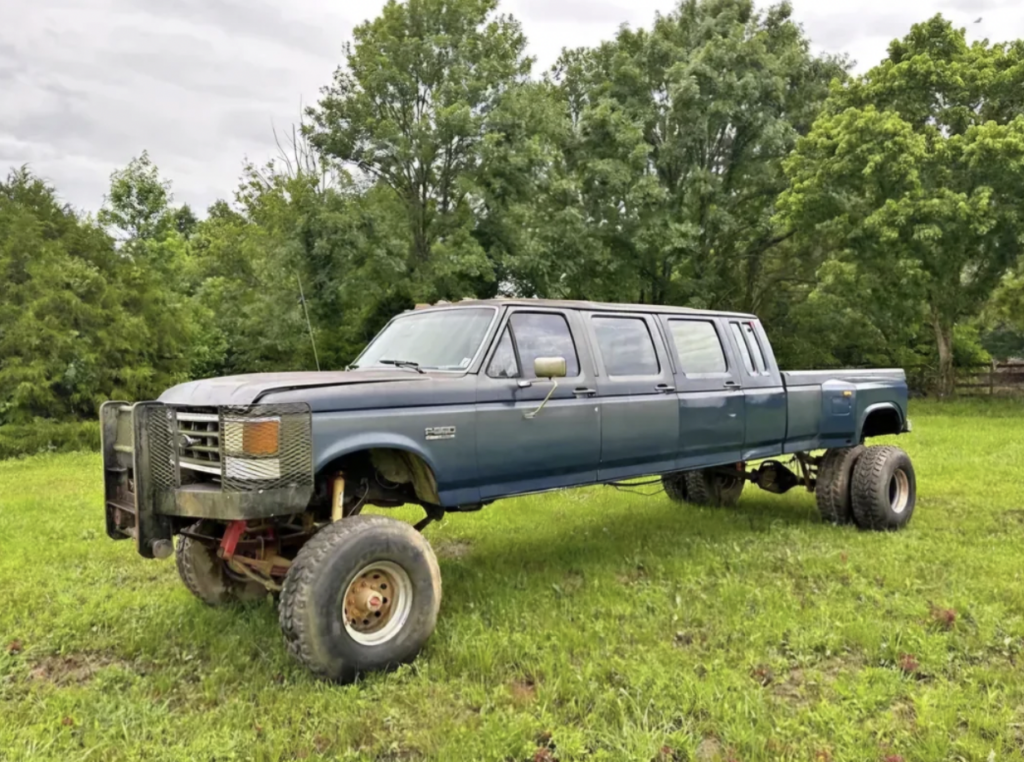 A customized, extra-long pickup truck with an elongated body, resembling a limousine, is parked on a grassy field. The truck has large off-road tires, a rugged front grille, and is painted in a weathered blue shade. Trees are visible in the background.