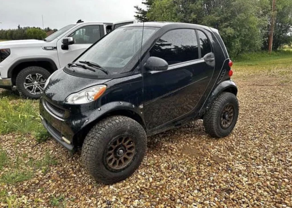 A black Smart car with large off-road tires and a lifted suspension is parked on a gravel surface. It is next to a white truck with trees and vegetation in the background.