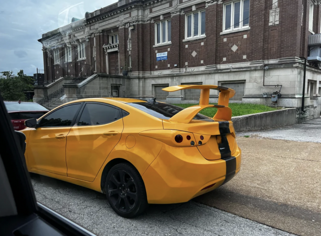 A bright yellow car with black accents, including a prominent rear spoiler and racing stripes, is parked on a street. The car is positioned next to a red vehicle, and a large brick building with arched windows serves as the backdrop.