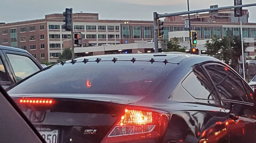 A black car with a spoiler adorned with diamond-shaped decorations is stopped at a traffic light in an urban area. Surrounding traffic signals and buildings are visible in the background. The car's tail lights and turn signals are illuminated.