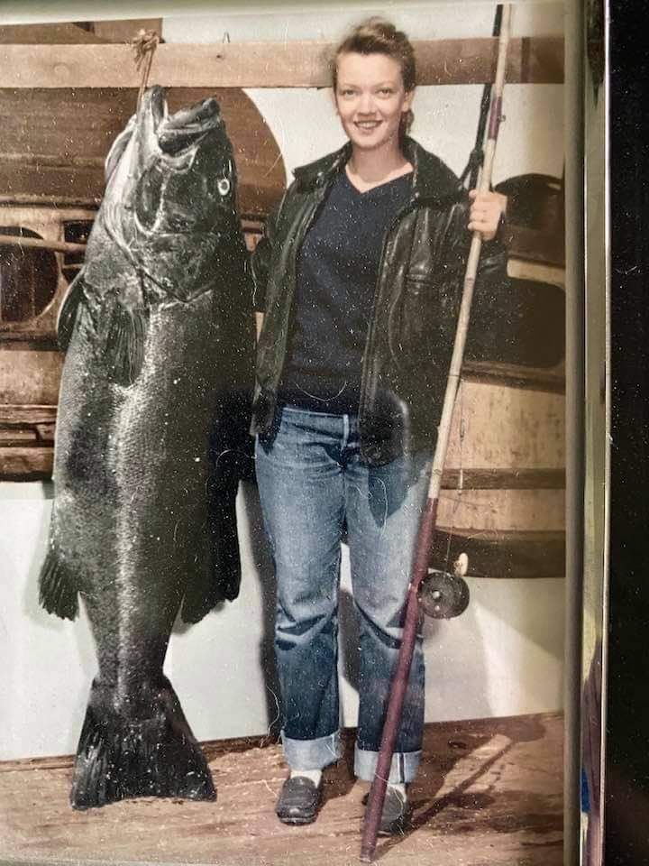 A woman stands on a boat holding a fishing rod with one hand and a large fish with the other. She is wearing a leather jacket, jeans, and sneakers, and she smiles at the camera. The fish is almost as tall as she is.