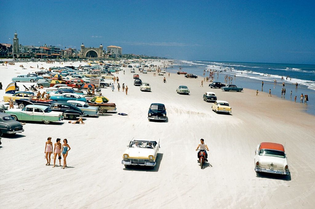 A crowded beach scene from the 1960s shows numerous vintage cars parked on the sand and people strolling or sunbathing. The ocean waves crash to the right while beachgoers enjoy the sunny day, and a motorcyclist rides along the beach amidst the classic cars.