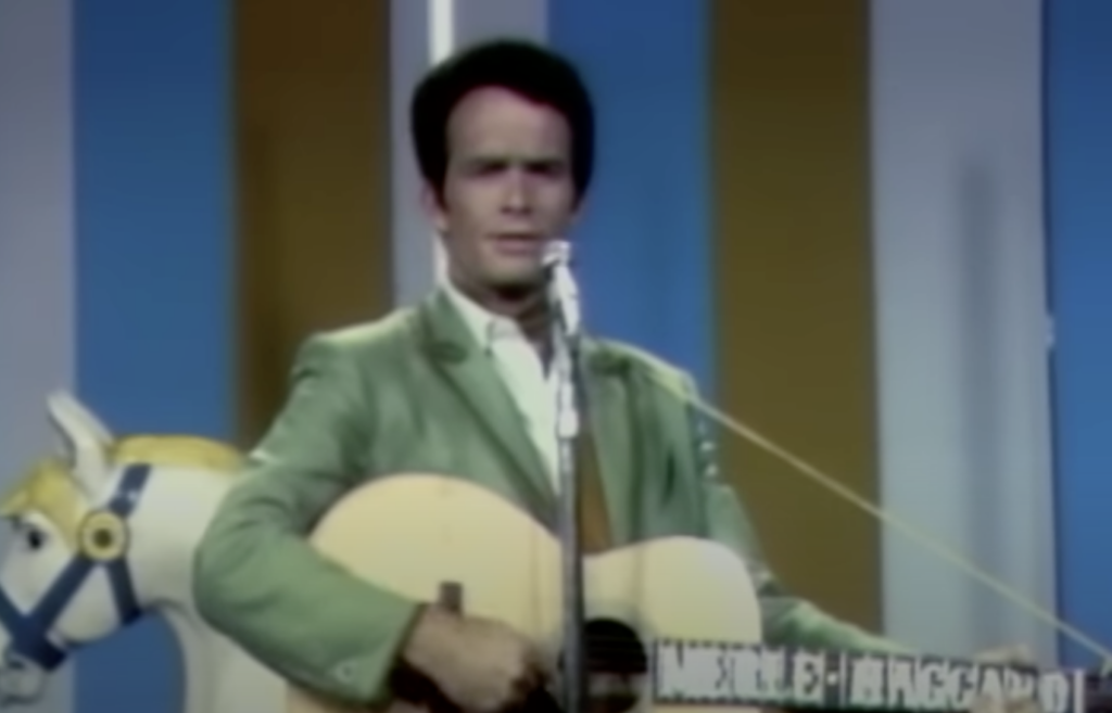 A man stands on stage, dressed in a light green jacket and white shirt, playing an acoustic guitar. Behind him is a prop horse, and the background displays vertical panels in white, blue, and yellow. He is singing into a microphone.