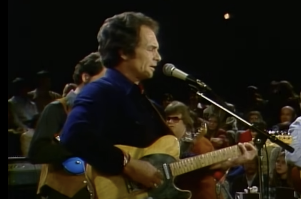 A man is performing on a stage, playing an electric guitar and singing into a microphone. He is wearing a dark shirt. The background shows other musicians playing instruments and an audience watching the performance. The lighting is dim, giving a warm ambiance.