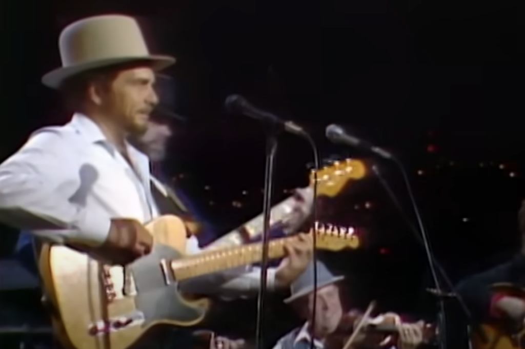 A man in a white hat and white shirt is playing an electric guitar on stage. He is singing into a microphone. Behind him are other musicians, including one playing a violin. The background is dark with twinkling lights.