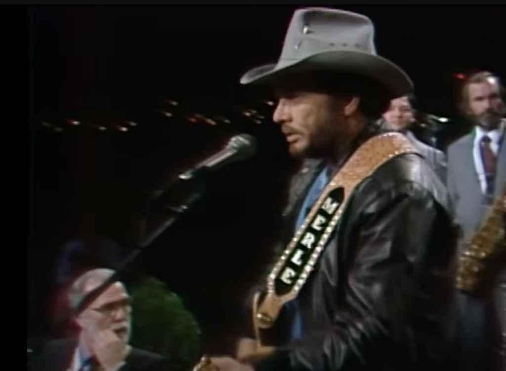 A musician wearing a cowboy hat and a leather jacket performs on stage. He holds an electric guitar with a strap bearing the name "Merle." In the background, a saxophonist and other band members are visible, along with stage lights and equipment.