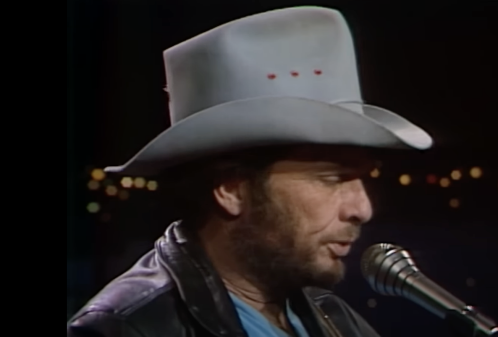 A person wearing a light grey cowboy hat and a black leather jacket is speaking or singing into a microphone. The background is dark with blurred lights.