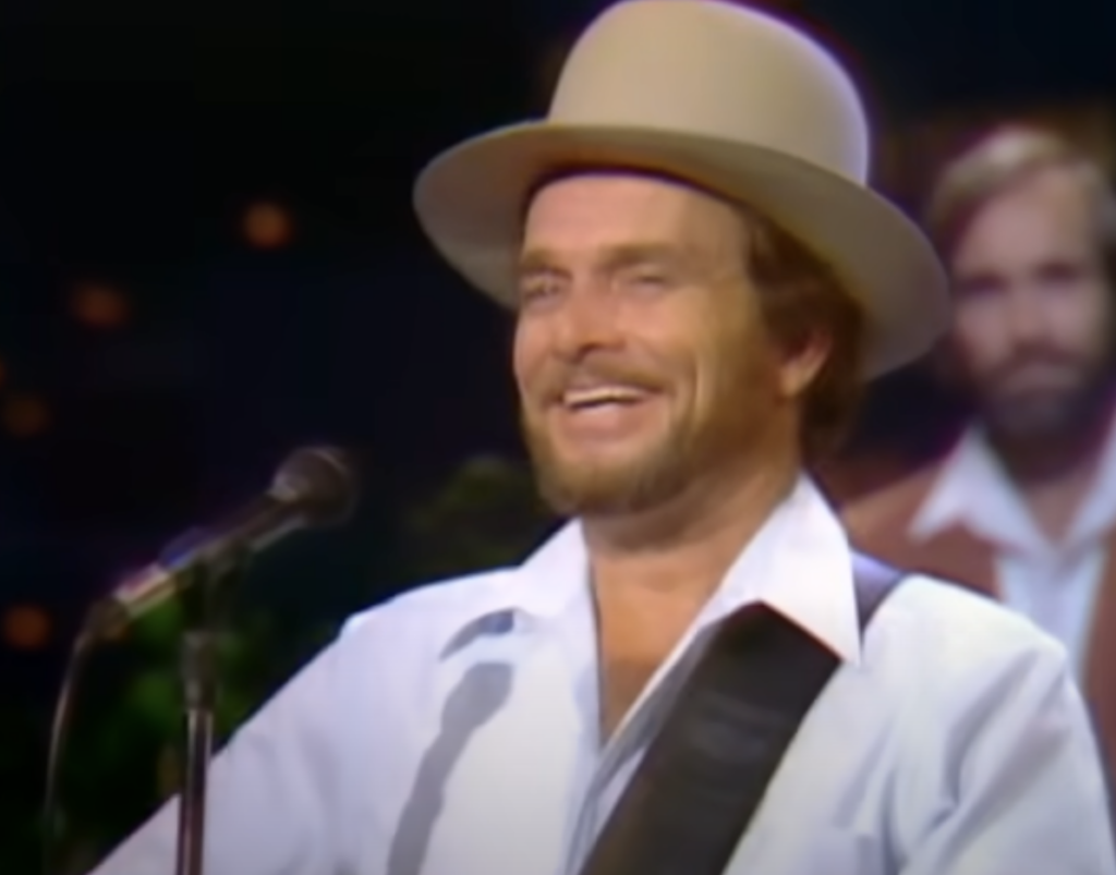 A man, wearing a light-colored cowboy hat and a white shirt with a guitar strap over his shoulder, is smiling as he stands in front of a microphone on stage. Another man in the background is slightly blurred and dressed in similar attire.