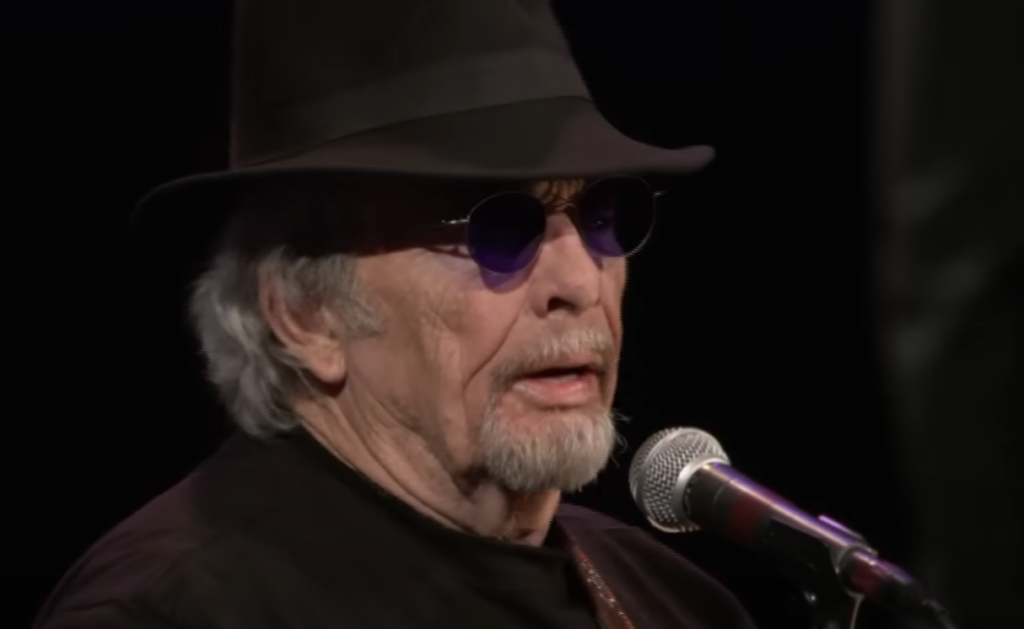 An older man with a gray beard and mustache is performing on stage. He is wearing a black hat, dark sunglasses, and a black outfit. A microphone is visible in front of him as he appears to be singing or speaking.