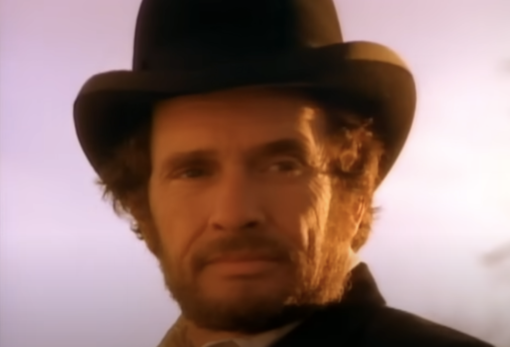 A bearded man wearing a black top hat and suit stands against a soft, dusky sky. His expression is contemplative, and the warm lighting highlights his features and the texture of his curly hair.