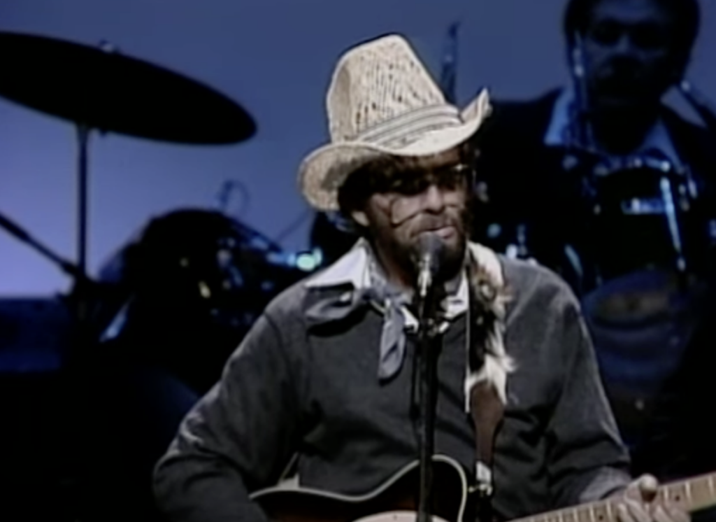 Person in a straw hat, sunglasses, and bandana around neck, playing guitar and singing into a microphone on stage. An out-of-focus drummer is visible in the background.