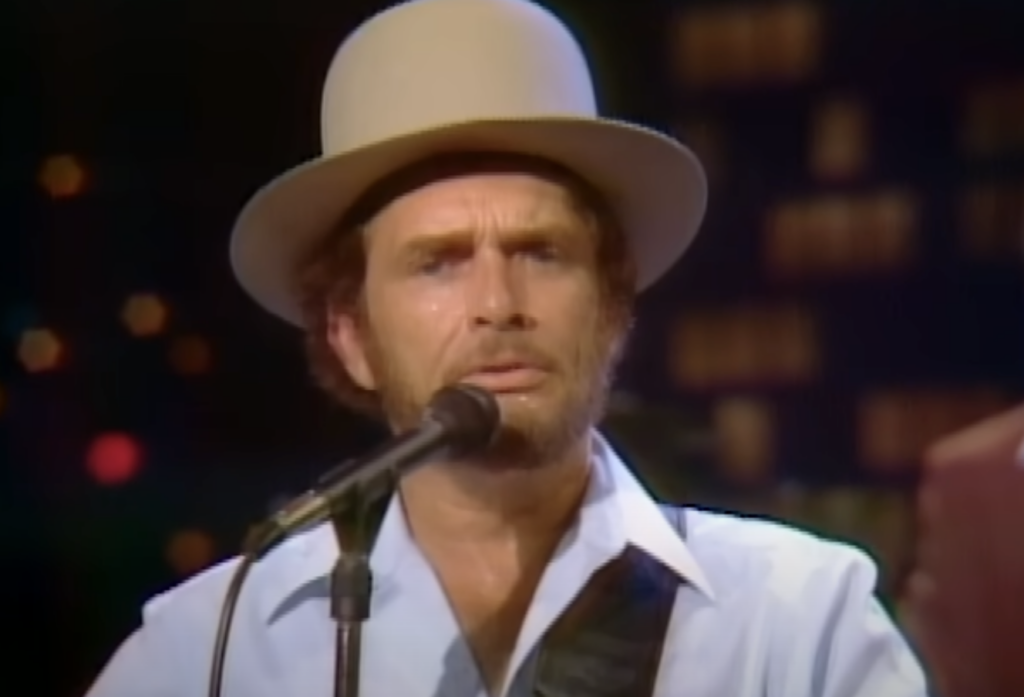 A man with a beard and mustache is performing on stage. He is wearing a light-colored hat and a light blue shirt, and he appears to be singing into a microphone. The background is dimly lit with colorful lights, creating a bokeh effect.