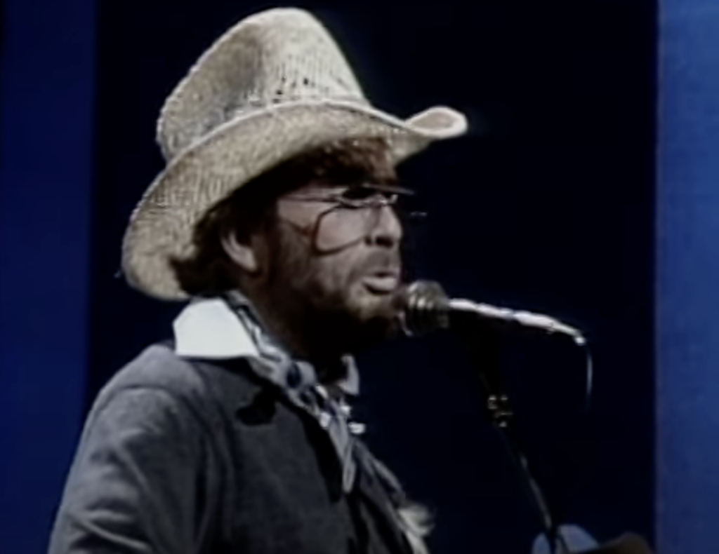 A person wearing a cowboy hat and glasses sings into a microphone. They have a beard and are dressed in a dark outfit with a light-colored scarf. The background is a gradient of dark blue to black.