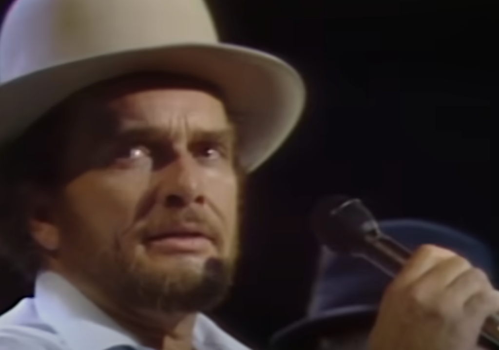 A man with a beard is wearing a cream-colored hat and holding a microphone close to his mouth while looking slightly to the side. The background is dark, suggesting a performance setting.